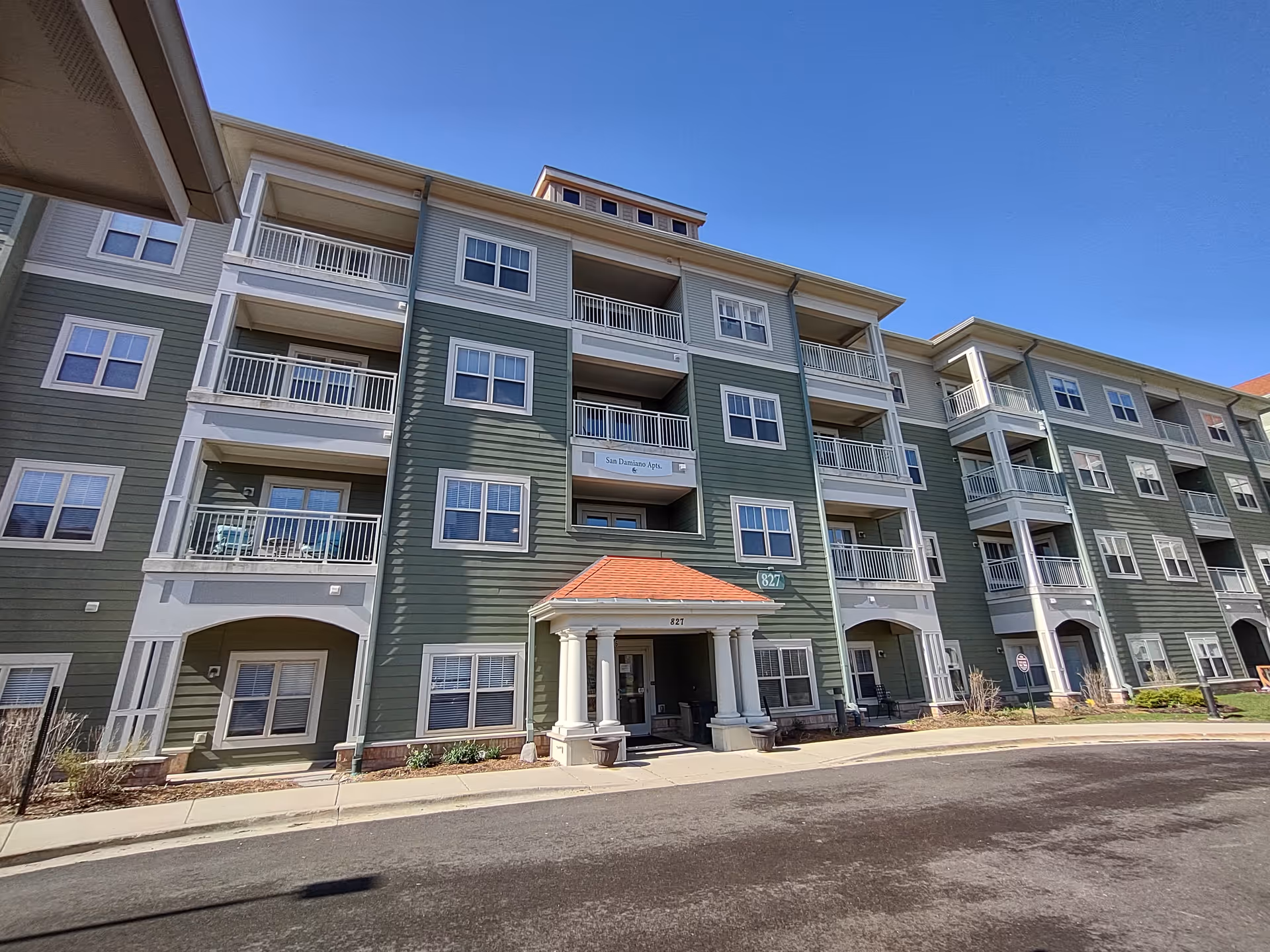 Exterior view of a multi-story residential building with green siding and white trim, featuring balconies and a covered entrance with columns under a clear blue sky.