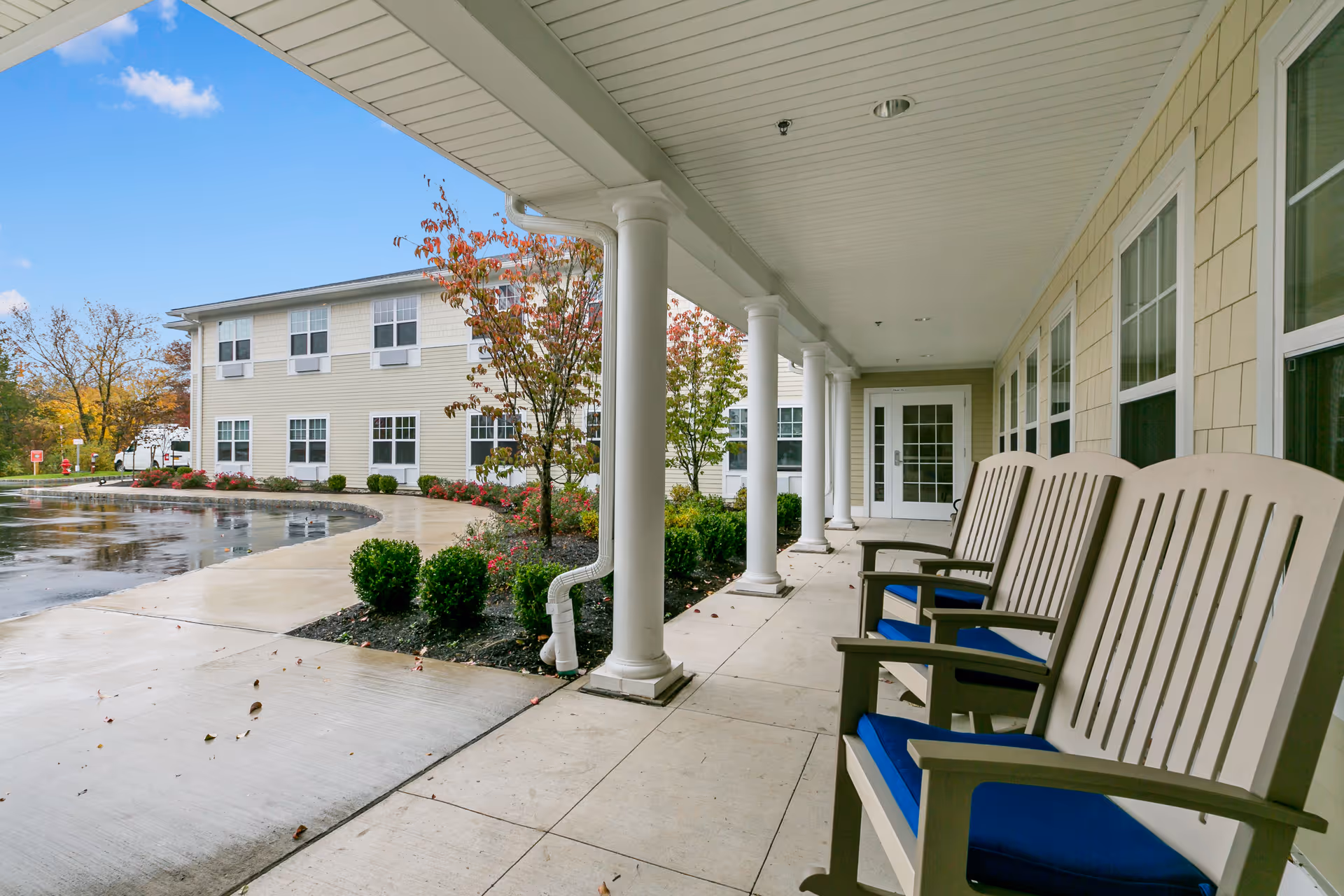 Covered front porch with columns and blue-cushioned chairs facing the entrance of a senior living building.