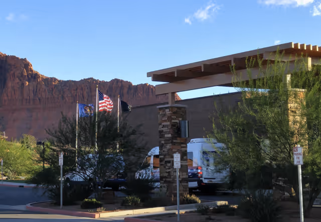 Exterior view of Southern Utah Veterans Home – Ivins with a covered entrance, several trees, three flagpoles displaying flags including the American flag, and a mountainous backdrop under a clear blue sky.