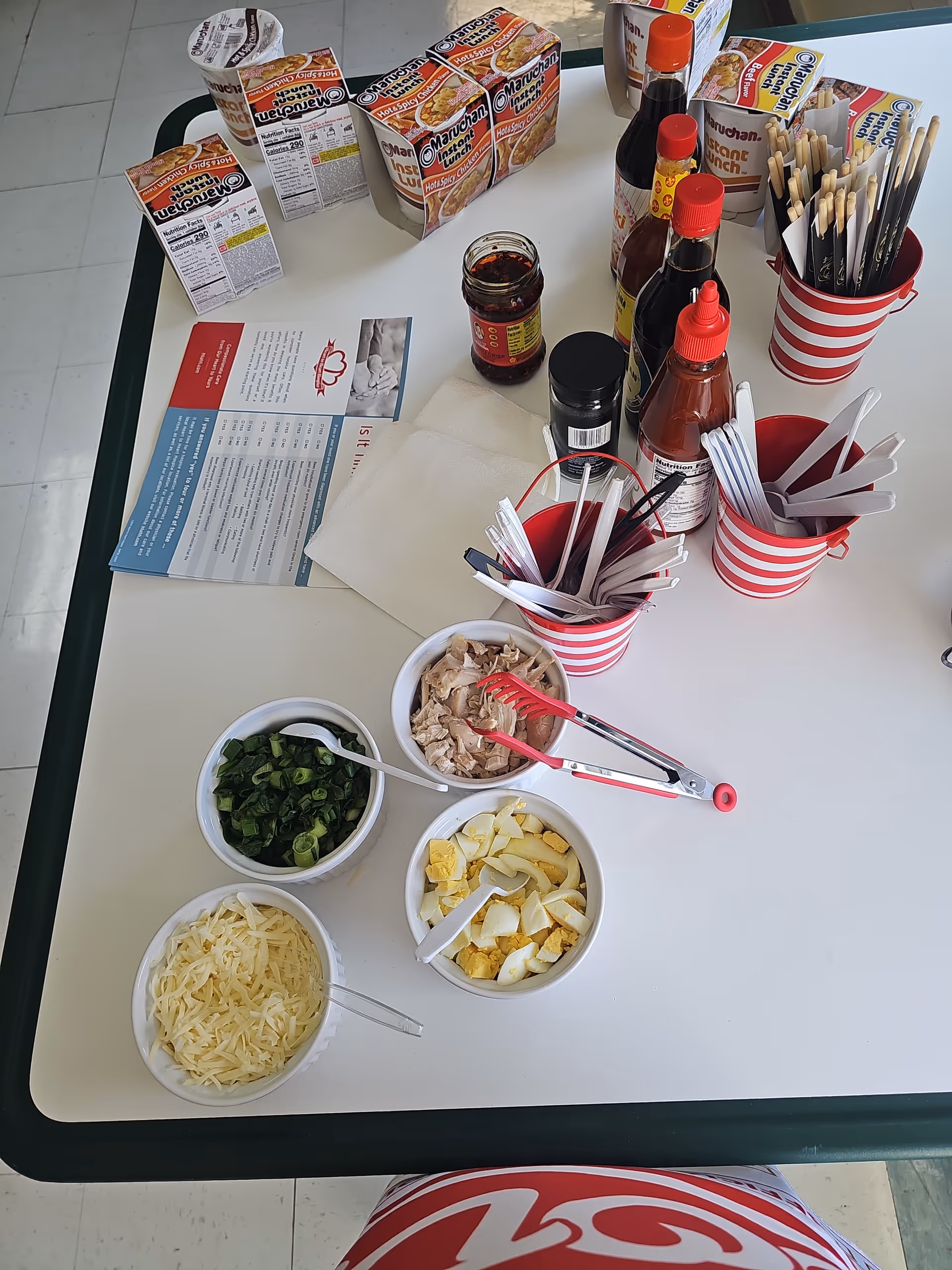 Table with condiments, utensils, napkins, and bowls of chopped ingredients in a dining area.
