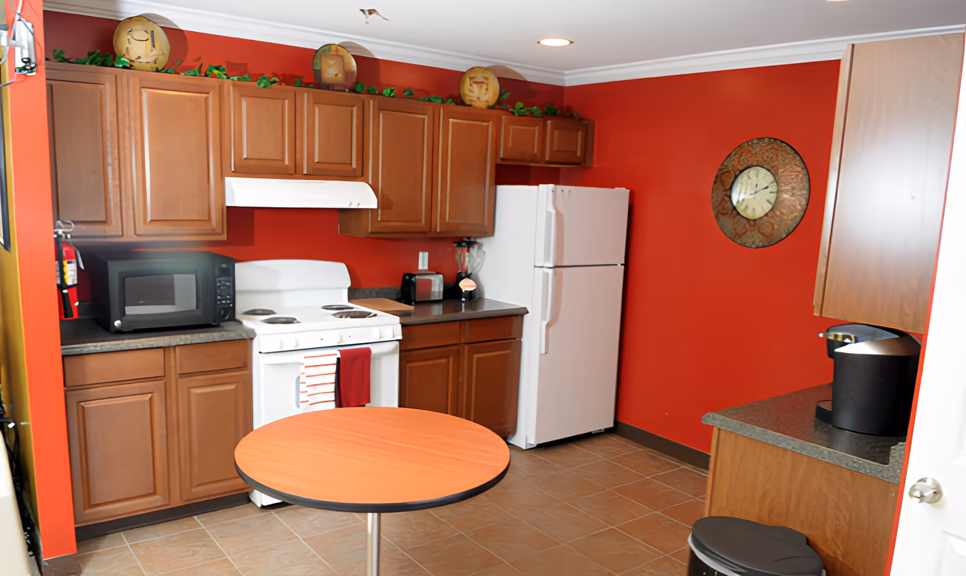 A kitchen with red walls, wooden cabinets, a white stove with a red and white towel hanging on the handle, a white refrigerator, a black microwave, a toaster, a blender, a coffee maker, a round wooden table, and a wall clock.