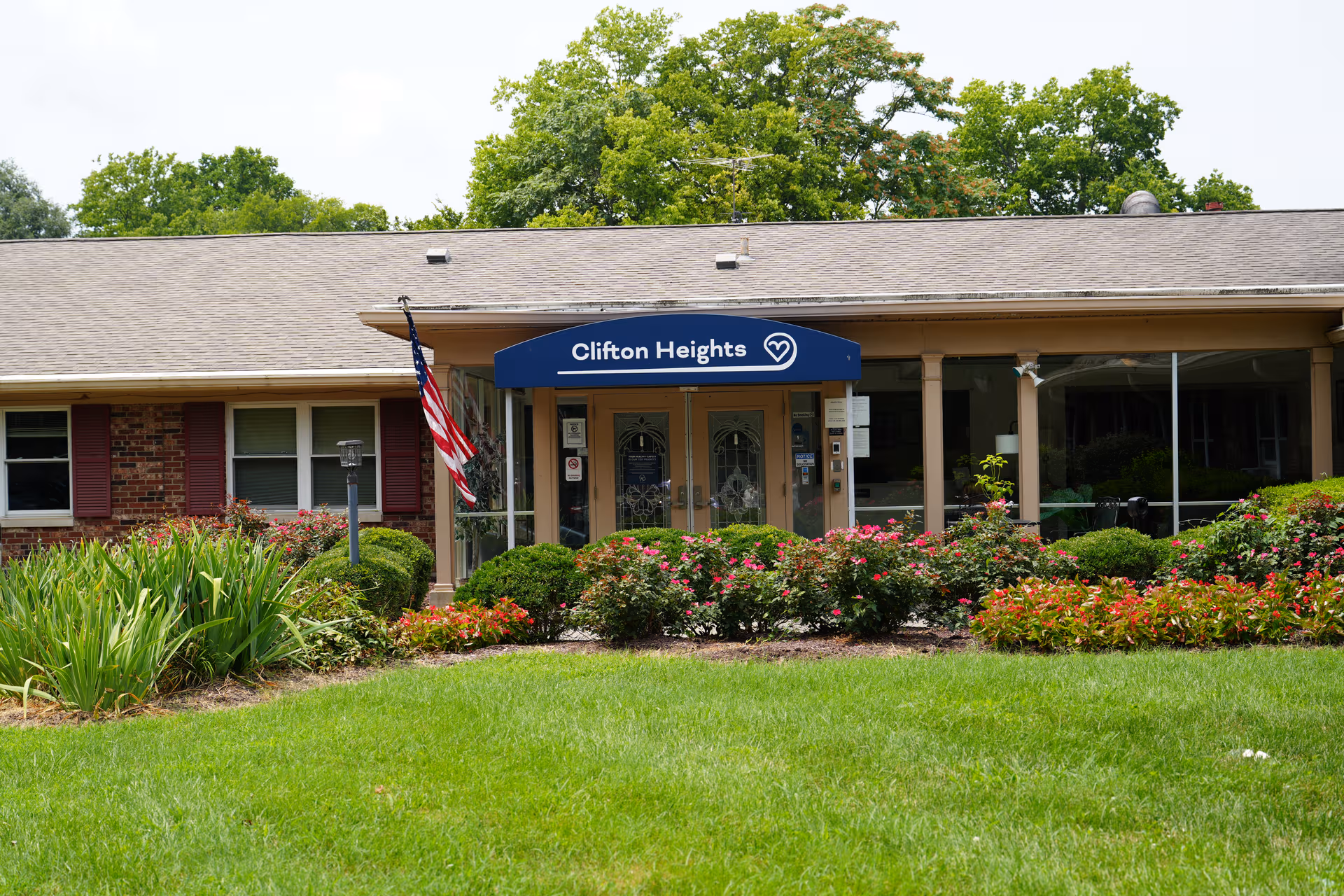 Front of Clifton Heights senior living facility with entrance, flag, and landscaped garden.