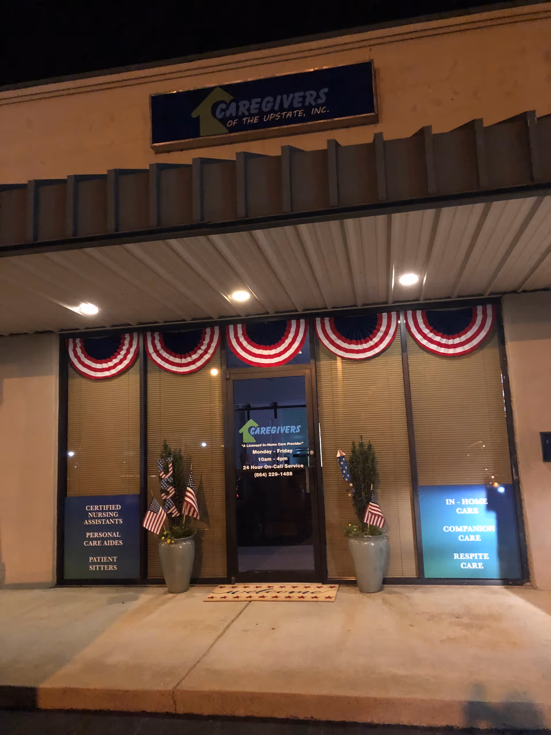 Night exterior of Caregivers of the Upstate storefront with patriotic bunting and potted plants.