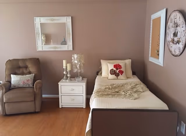 A cozy bedroom with a single bed covered in a cream-colored blanket and a decorative pillow with red flowers. Next to the bed is a white nightstand with two drawers, holding two candles and a small decorative lamp. To the left of the nightstand is a brown cushioned armchair with a floral pillow. The walls are painted light brown and decorated with a white-framed mirror, a white-framed corkboard, and a large round clock.