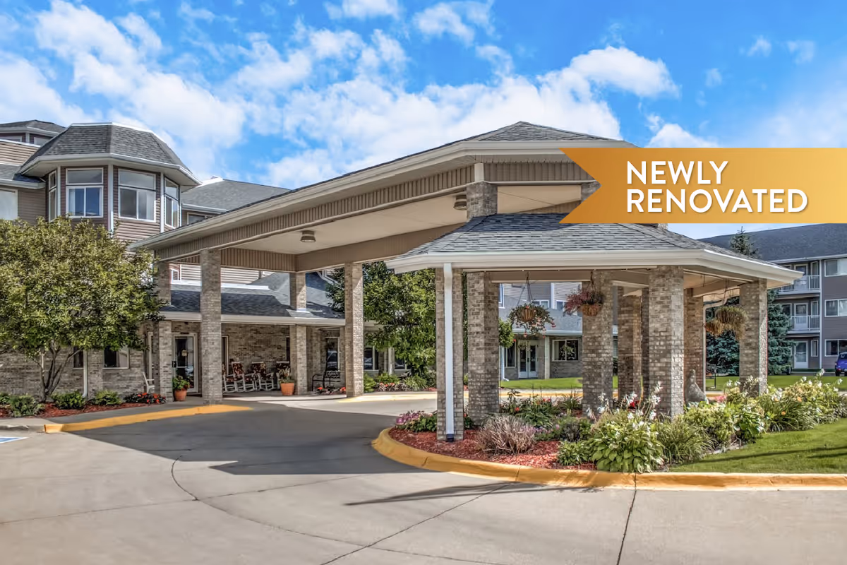 Exterior view of Mallard Point Senior Living showing a covered entrance with brick pillars, landscaped garden beds with flowers and shrubs, and a clear blue sky with some clouds. A banner in the top right corner reads 'NEWLY RENOVATED'.