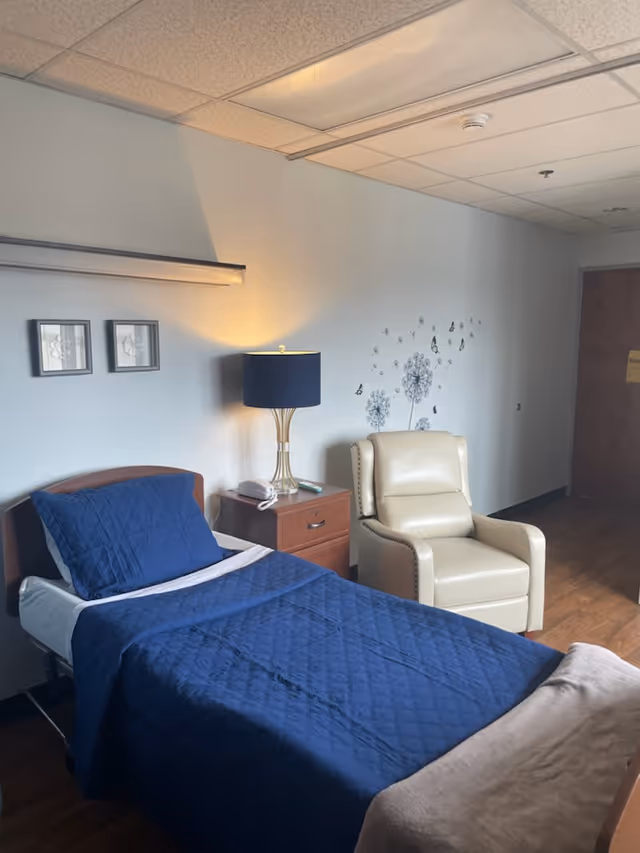 A blue quilted bed with pillows beside a beige recliner in a senior room.