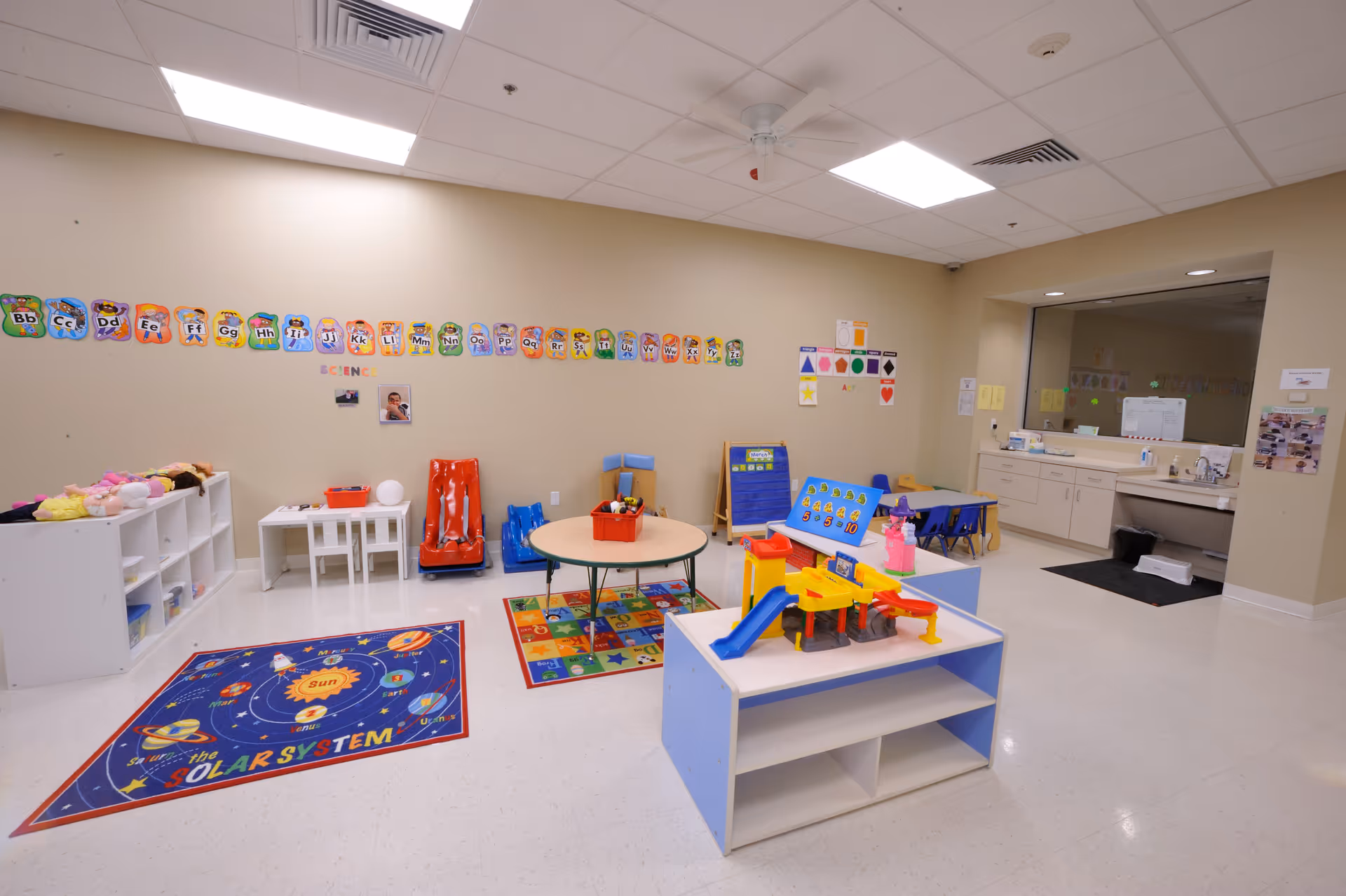 Bright indoor playroom with colorful toys, tables, cubbies, and a solar-system rug.