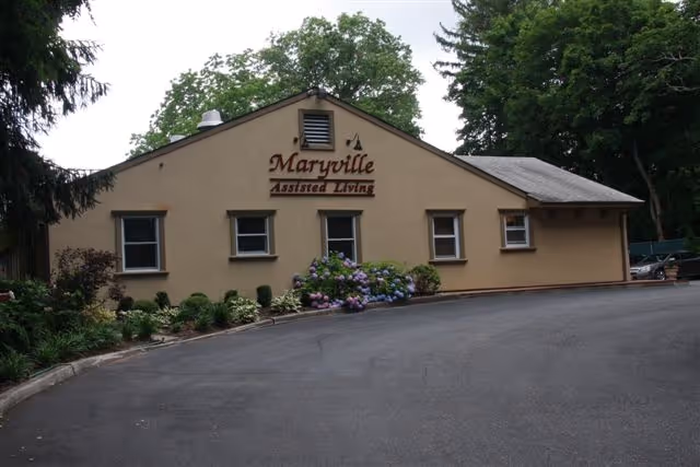 Front exterior of a single-story beige building reading "Maryville Assisted Living" with shrubs and a curved driveway.