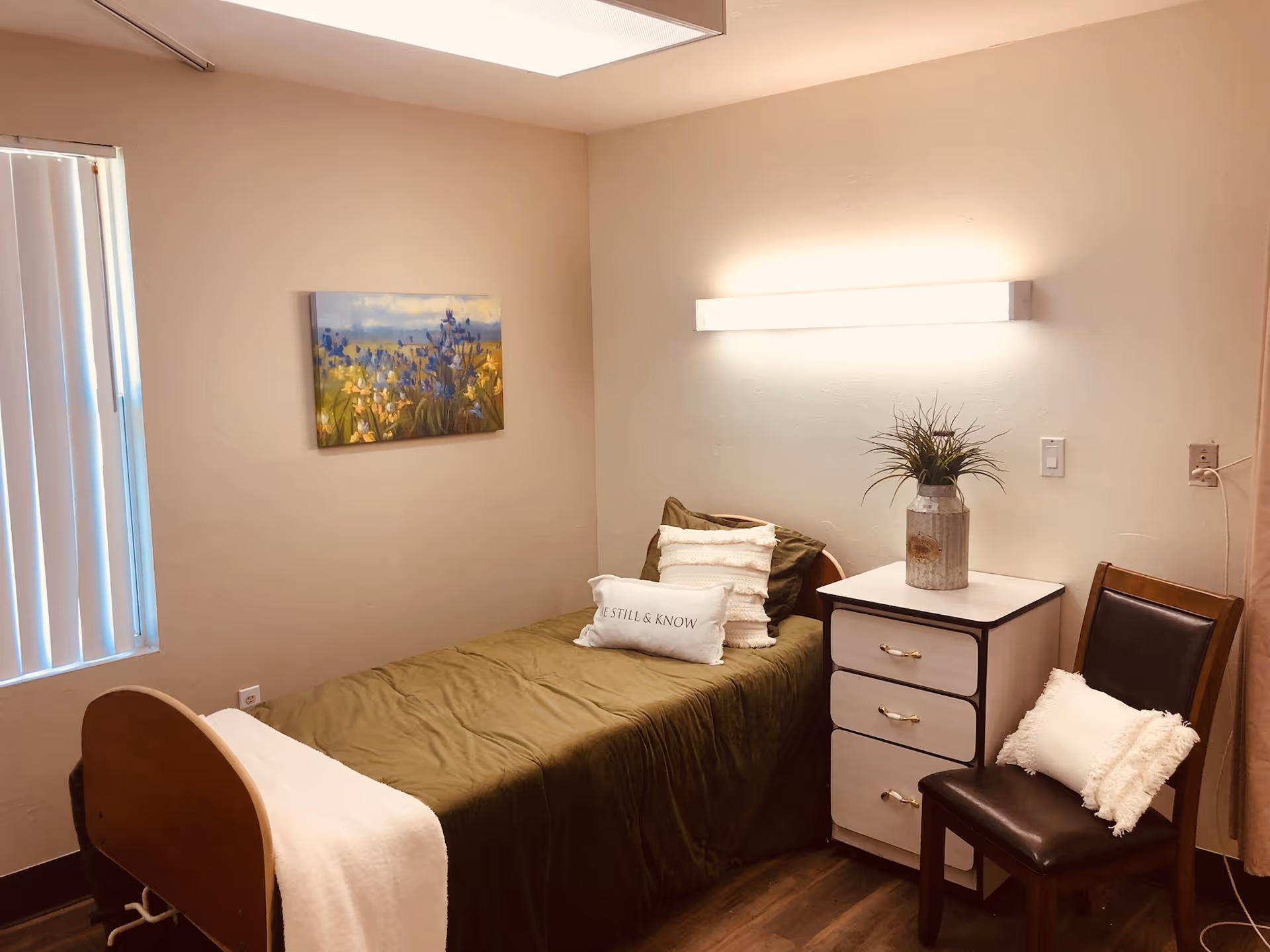 Small furnished single-occupancy bedroom with a bed, nightstand, chair, wall art, and vertical blinds.