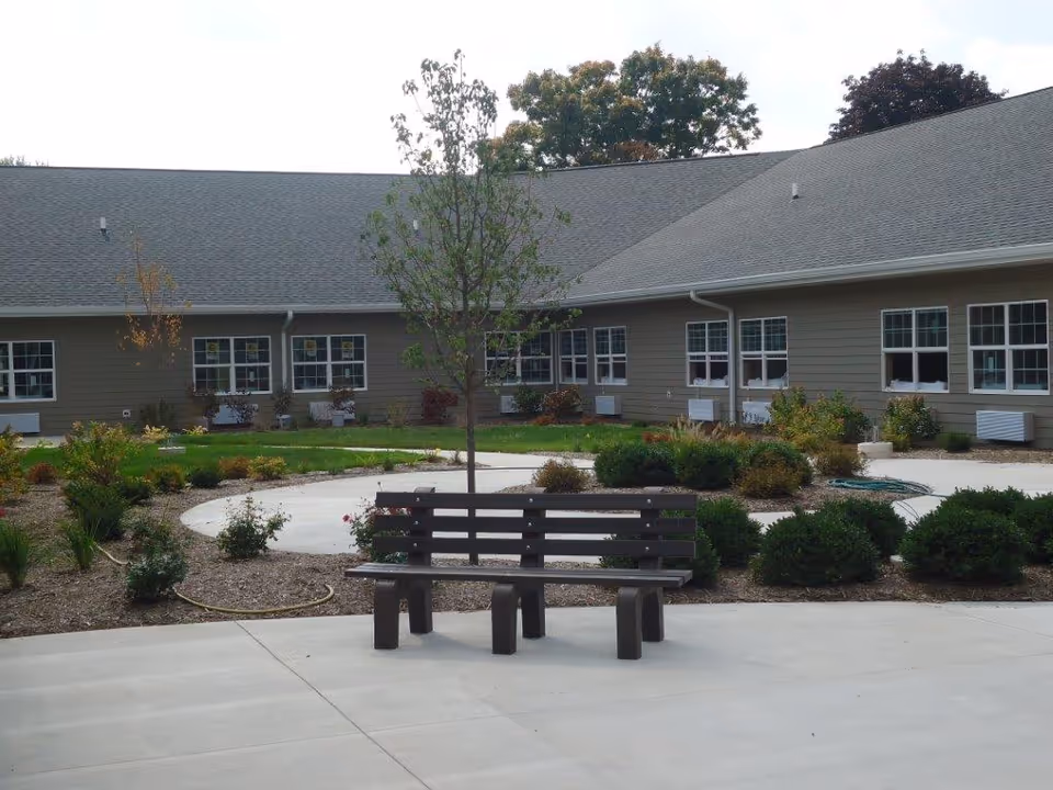 Outdoor courtyard area of a senior living facility with a wooden bench in the foreground, a young tree planted behind it, surrounded by landscaped bushes and plants, and a building with multiple windows in the background.