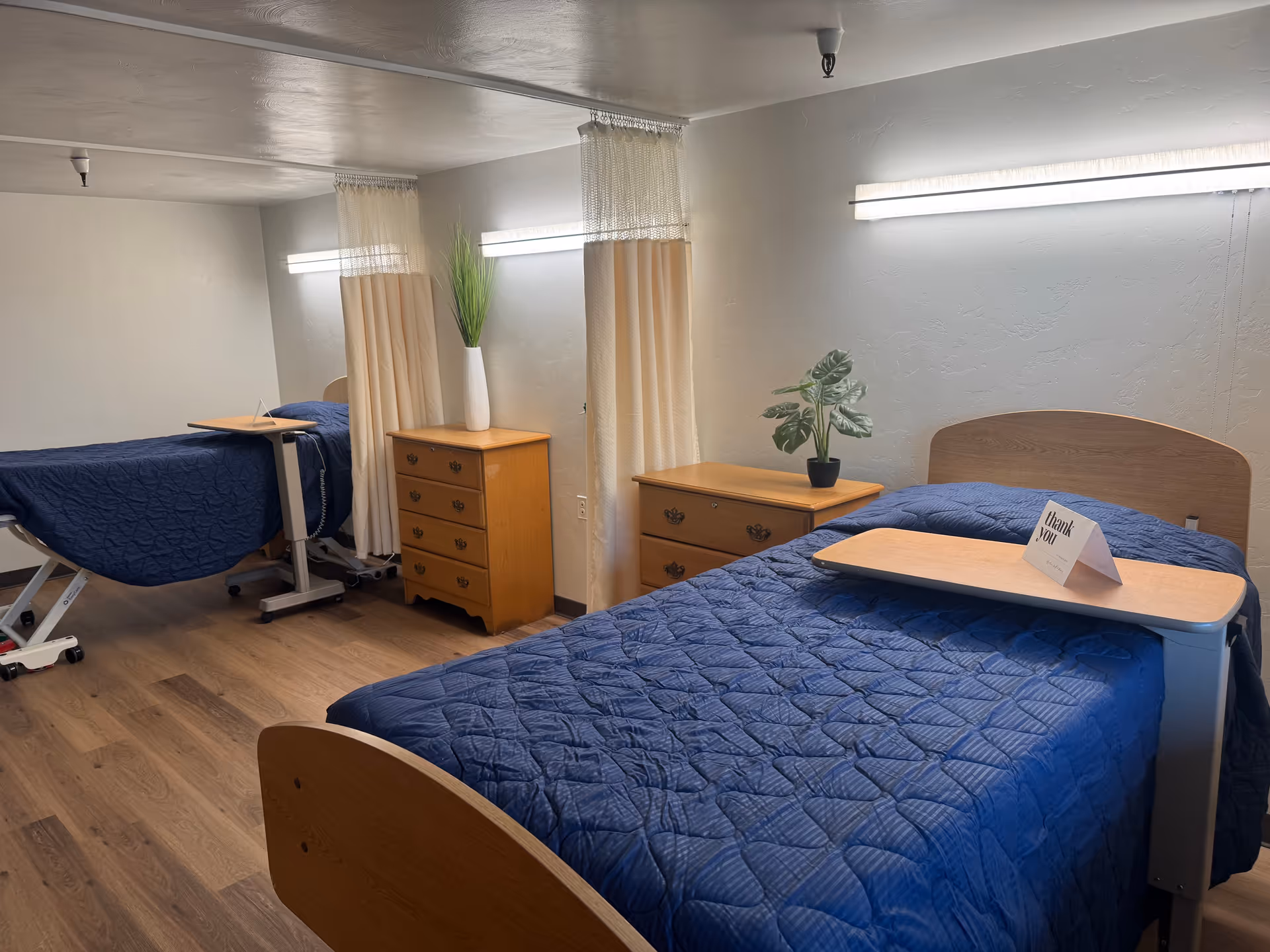 Two single beds with blue quilts, wooden dressers, and privacy curtains in a shared care room.