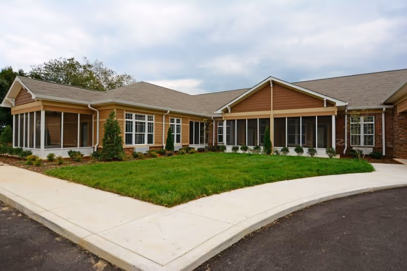 Exterior view of a single-story building with brown and beige siding, multiple windows, and a well-maintained green lawn bordered by a concrete sidewalk and driveway under a cloudy sky.