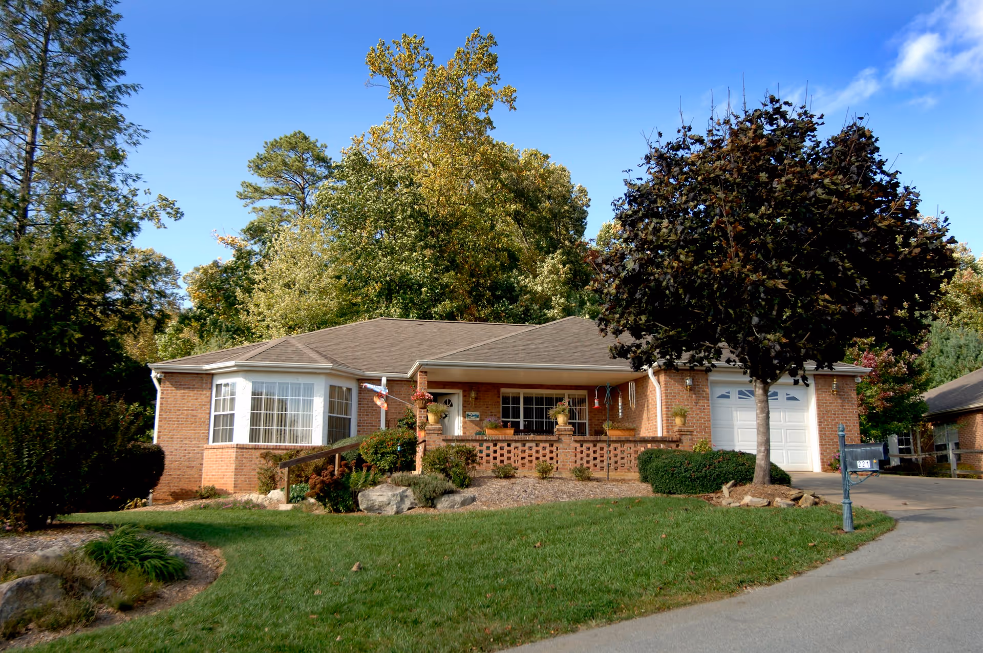 Single-story brick building with a front porch, large windows, a garage, and a well-maintained lawn with trees and shrubs under a clear blue sky.