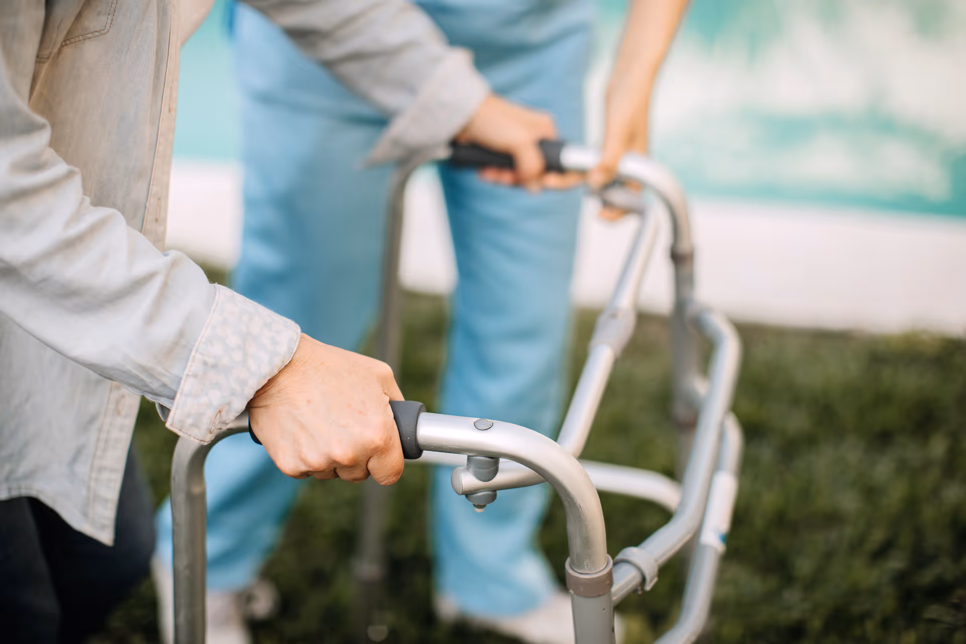 A caregiver's hand grips a mobility walker outdoors, with another caregiver in scrubs in the background.