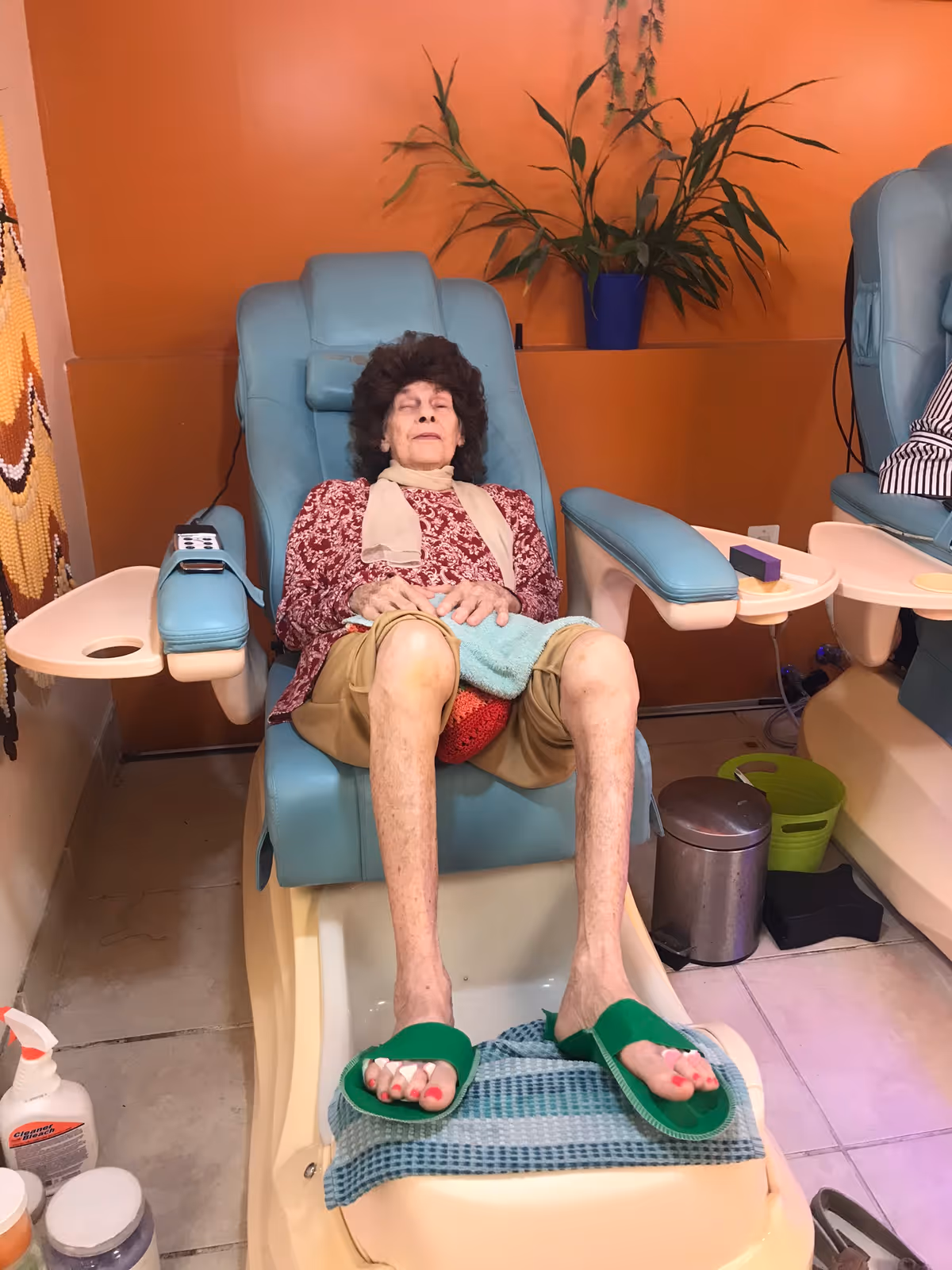 An elderly woman with curly hair is sitting in a blue pedicure chair with her eyes closed, wearing a red patterned top, beige shorts, and green slippers. Her toenails are painted red. The setting appears to be a salon or spa area with an orange wall, a potted plant in the background, and various salon supplies nearby.