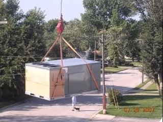 A large crate is hoisted by straps over a residential street with a person nearby.