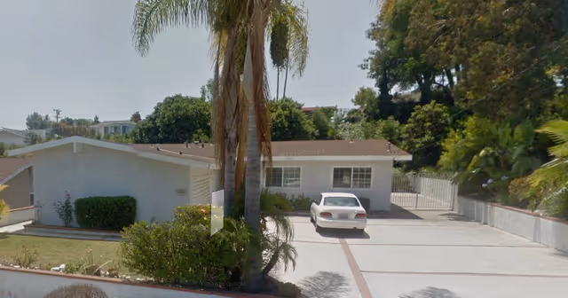 Front exterior of a single-story senior living building with driveway, palm trees, and a parked car.