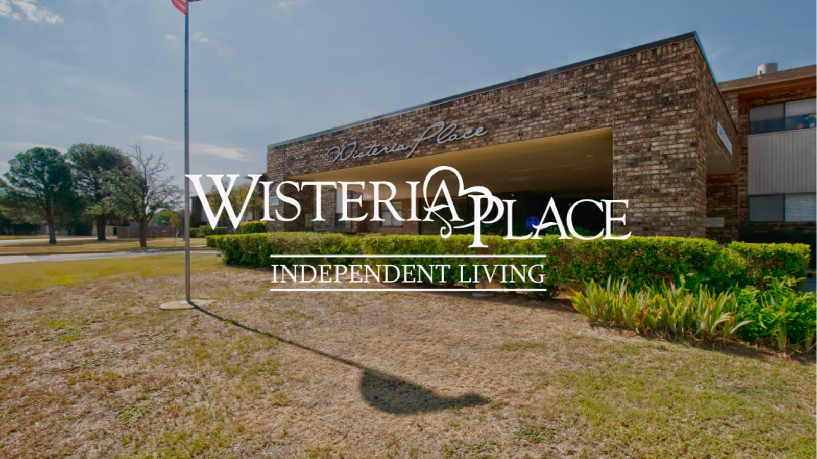 Exterior view of Wisteria Place Independent Living facility showing a brick building with a covered entrance, surrounded by grass, shrubs, and trees under a partly cloudy sky.