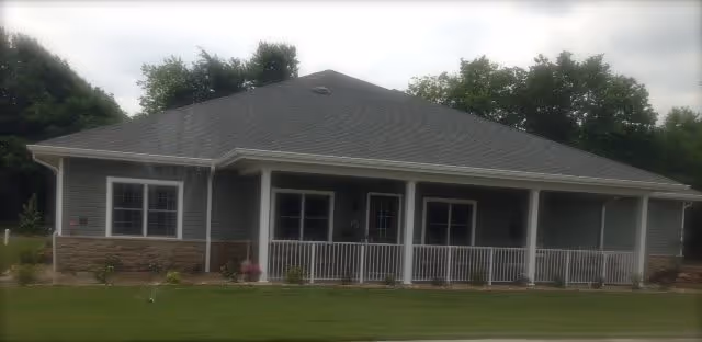 Single-story building with a gray roof, light gray siding, and stone accents along the lower exterior walls. The building has a covered porch with white railings and several windows. It is surrounded by a well-maintained lawn and trees in the background under a cloudy sky.