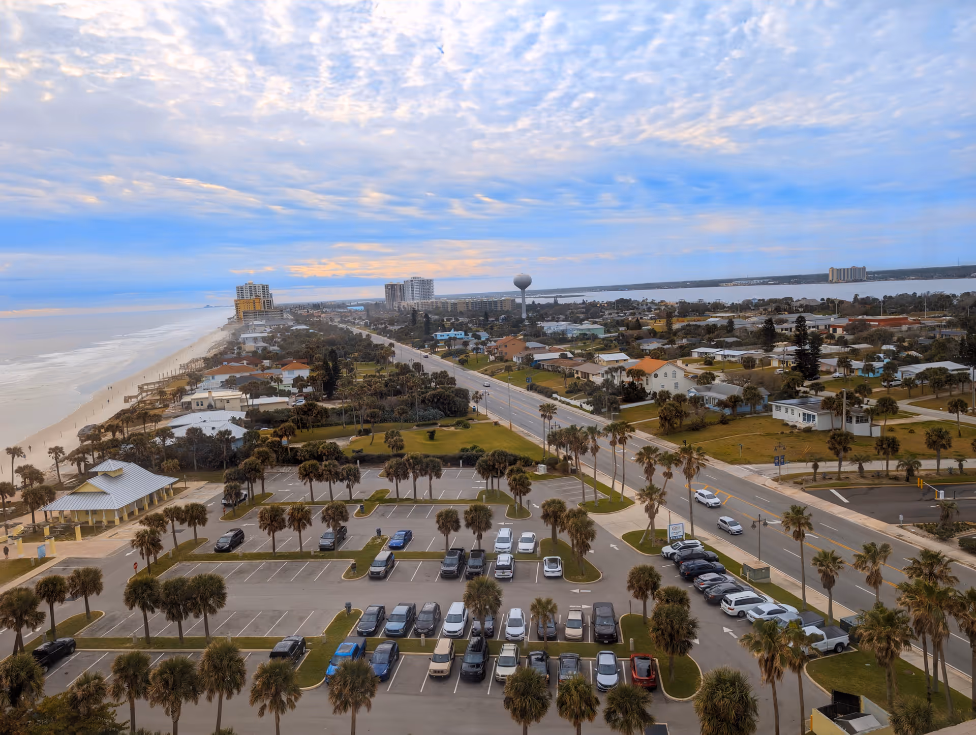 Aerial view of a coastal town with beach, roads, palm trees, and parking lots.