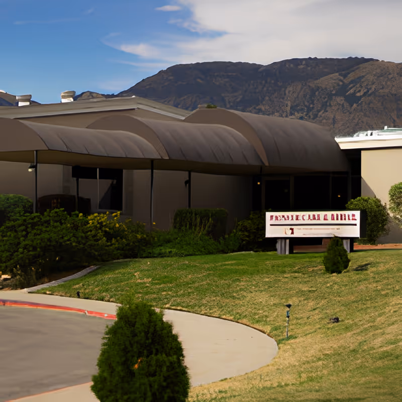 Front exterior entrance of a single-story building with a curved fabric awning, a lawn and shrubs, and mountains in the background.