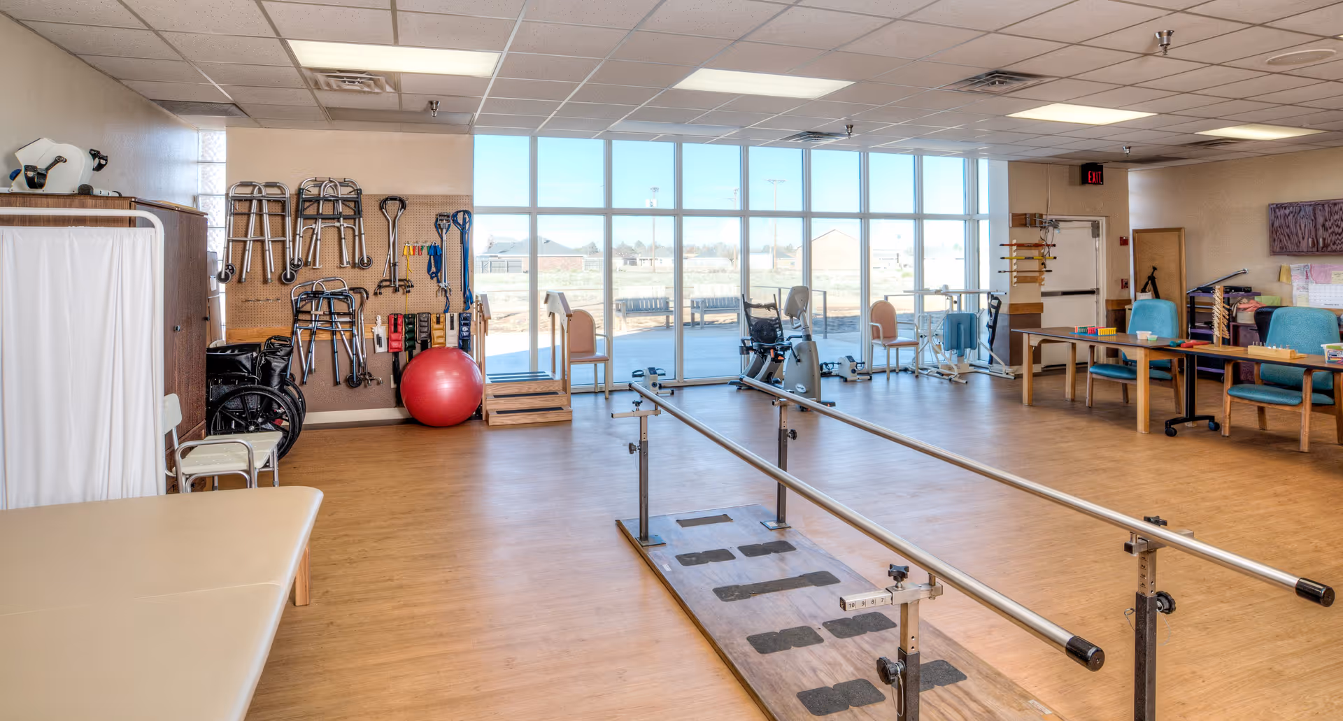 A spacious rehabilitation therapy room with parallel bars for walking exercises in the center. The room has large windows letting in natural light, various exercise equipment including walkers, wheelchairs, a red exercise ball, and therapy tables. There are chairs and tables along the right side of the room, and the floor is wooden.