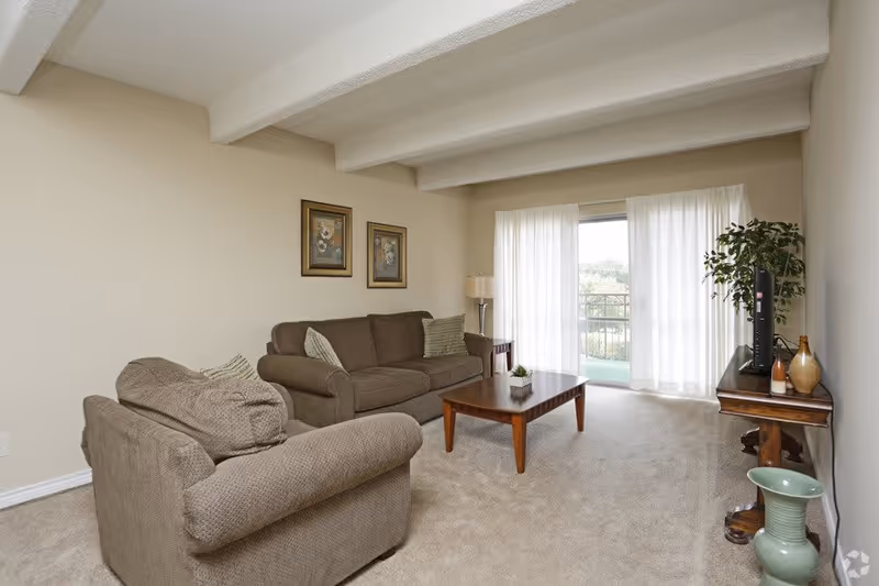 A cozy living room with beige walls and carpet, featuring a brown sofa and armchair with green striped pillows, a wooden coffee table, a side table with a lamp, a TV on a wooden stand, a potted plant, and large sliding glass doors with sheer white curtains letting in natural light.