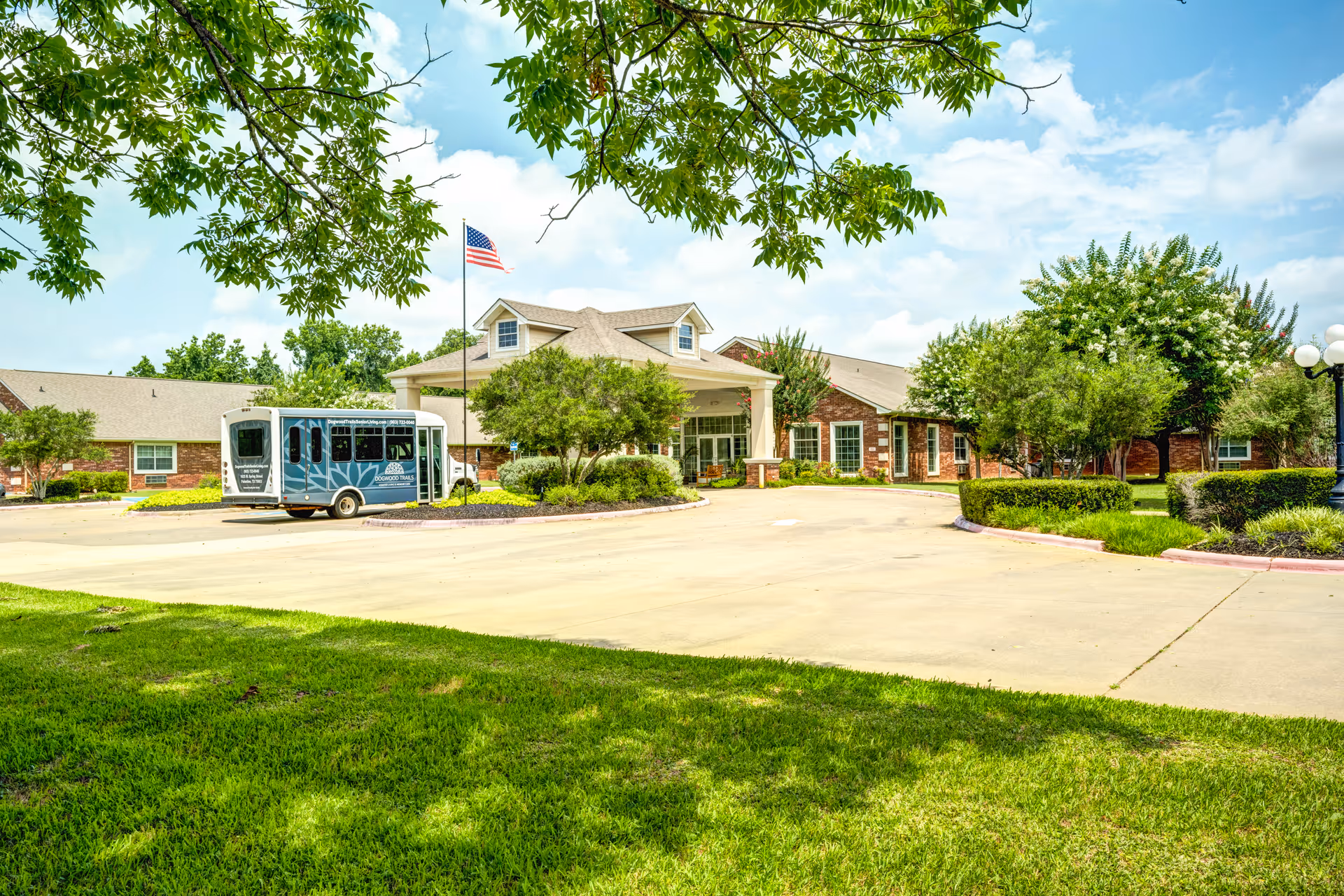 Wide view of Dogwood Trails Assisted Living exterior with entrance, landscaping, and a parked shuttle bus