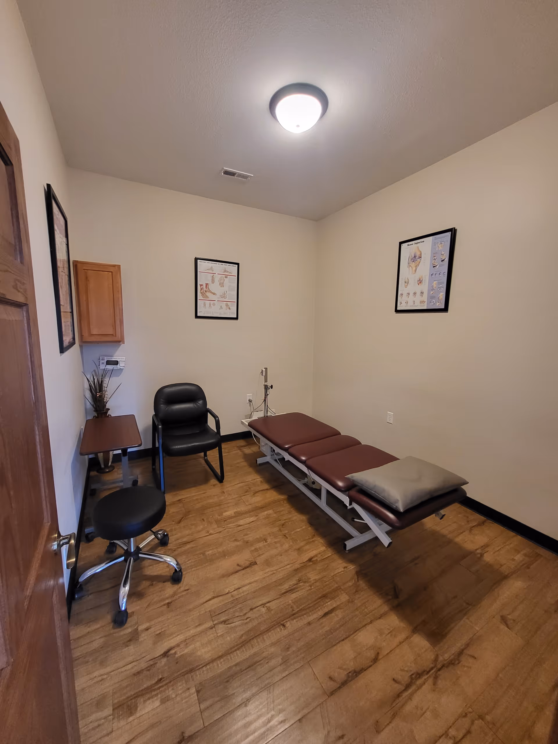 Small clinical examination room with a brown padded exam table, chairs, and wall charts.