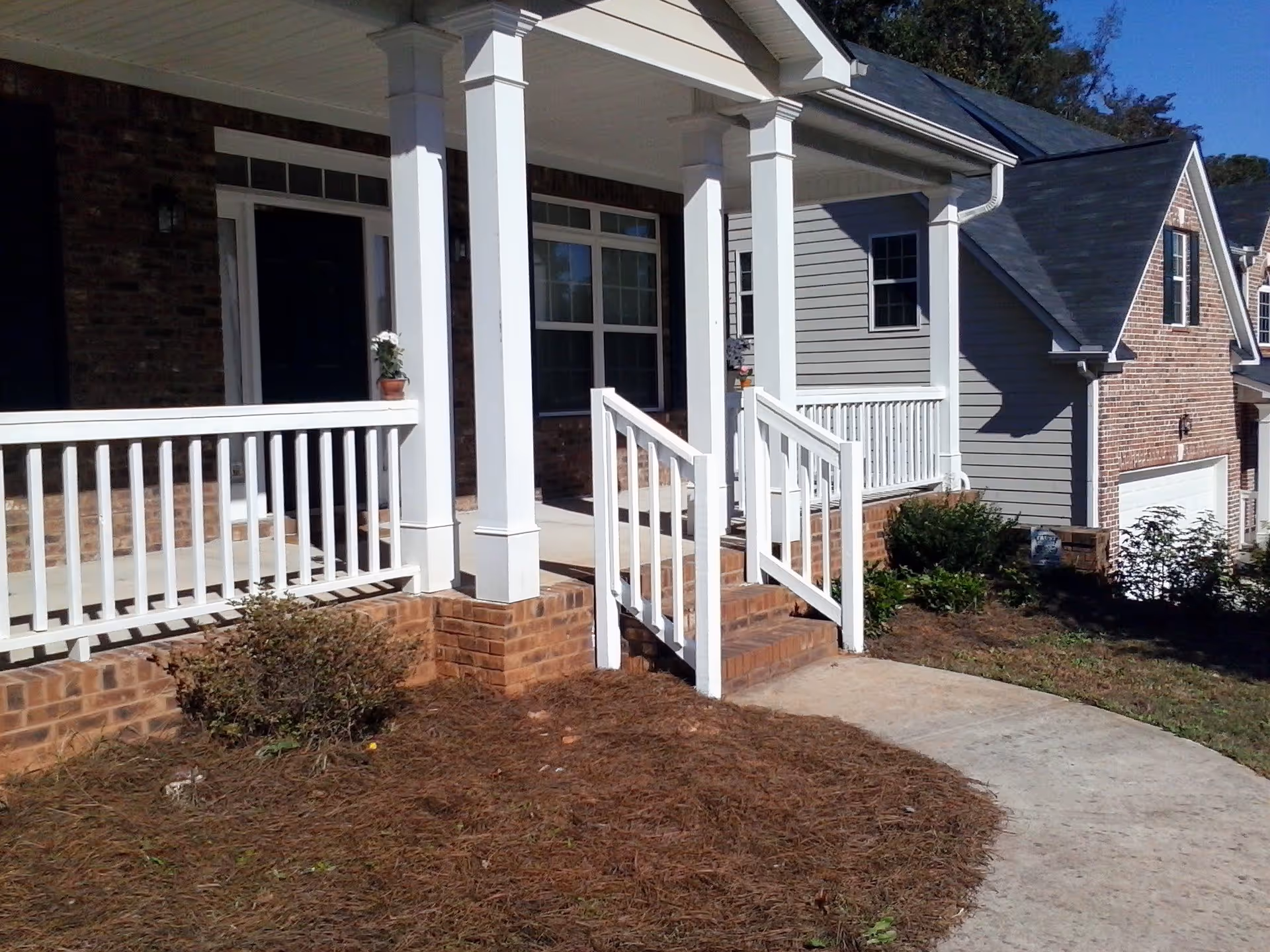 Front porch of a residential building with white railings and columns, brick steps, and a concrete walkway leading up to the entrance. There are shrubs and a small garden area around the porch.