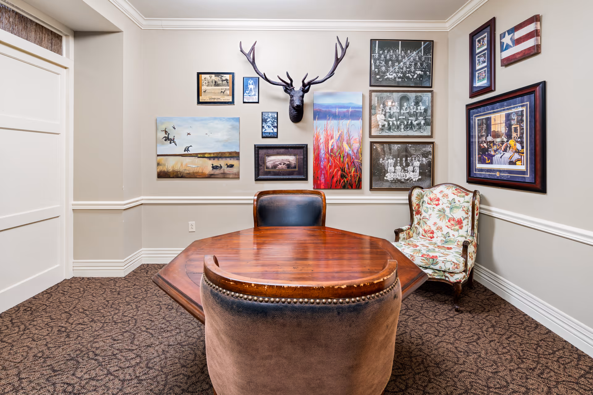 A small meeting or sitting room with a wooden table and three chairs, one upholstered in floral fabric and two with leather backs. The walls are decorated with various framed pictures, including black and white photos, paintings, and a mounted deer head. The room has beige walls with white trim and patterned carpet flooring.