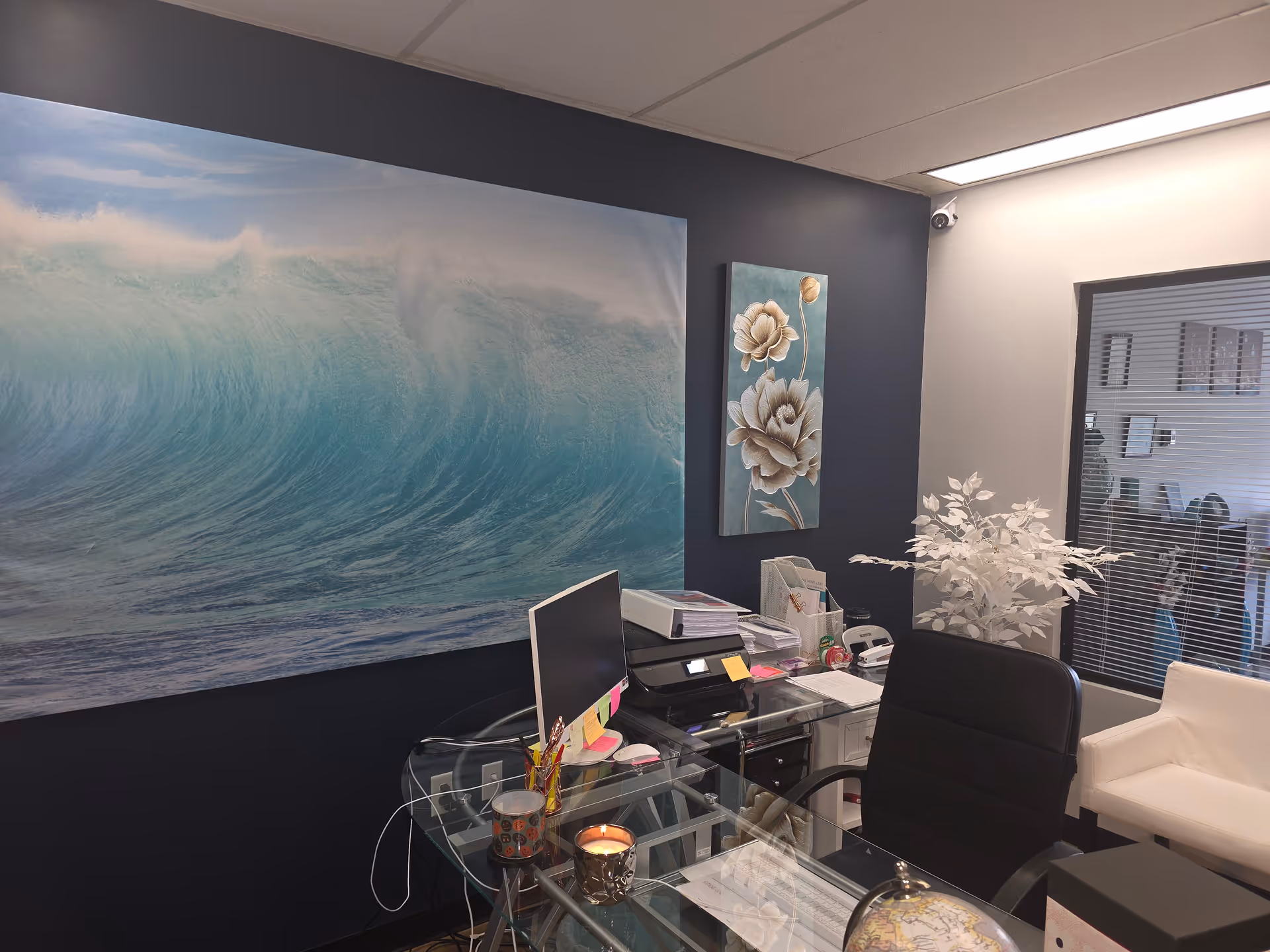 Glass desk with computer, printer, papers, and ocean-themed wall art in an office.