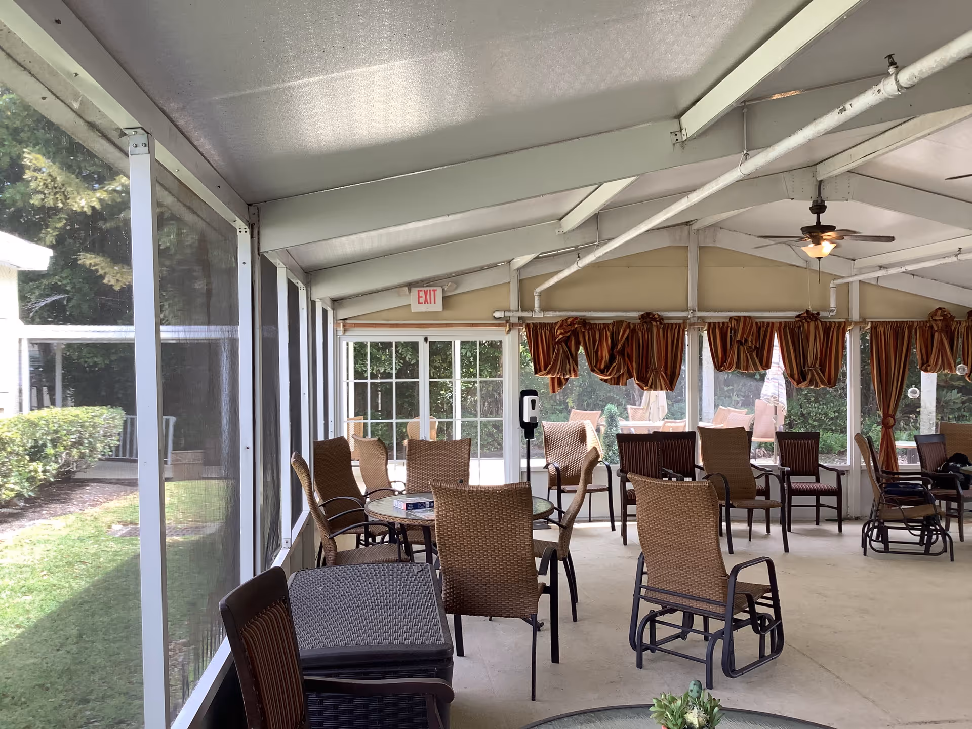 Covered outdoor seating area with wicker chairs and tables, glass walls, and ceiling fan.