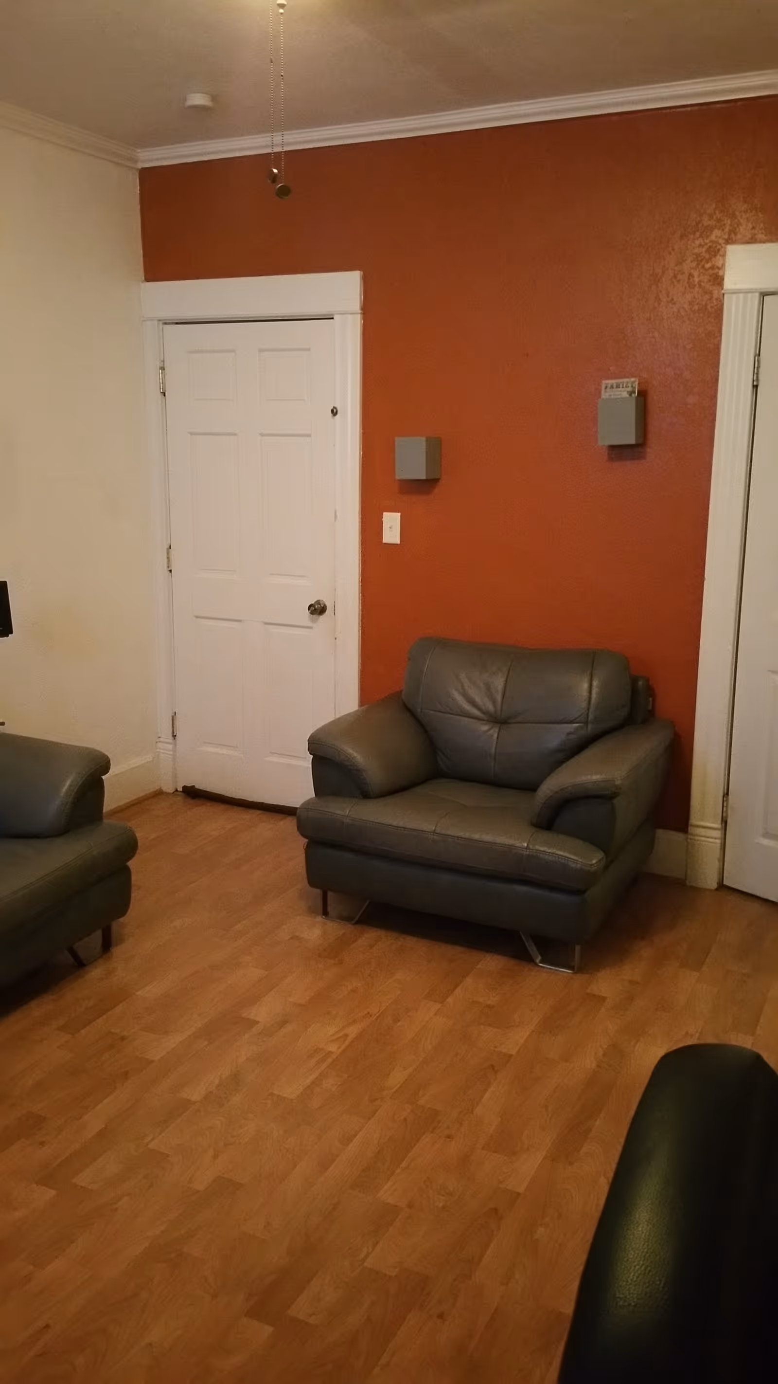 A living room with a gray leather armchair against an orange accent wall and a white door on hardwood floors.