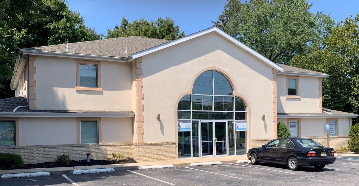 Beige two-story building with arched glass entrance and a parking lot in front.