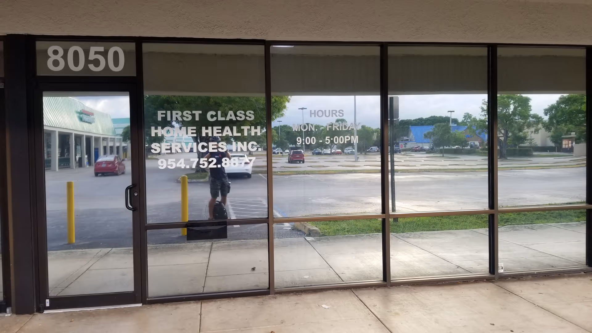 Glass storefront showing First Class Home Health Services sign and a reflection of the photographer.
