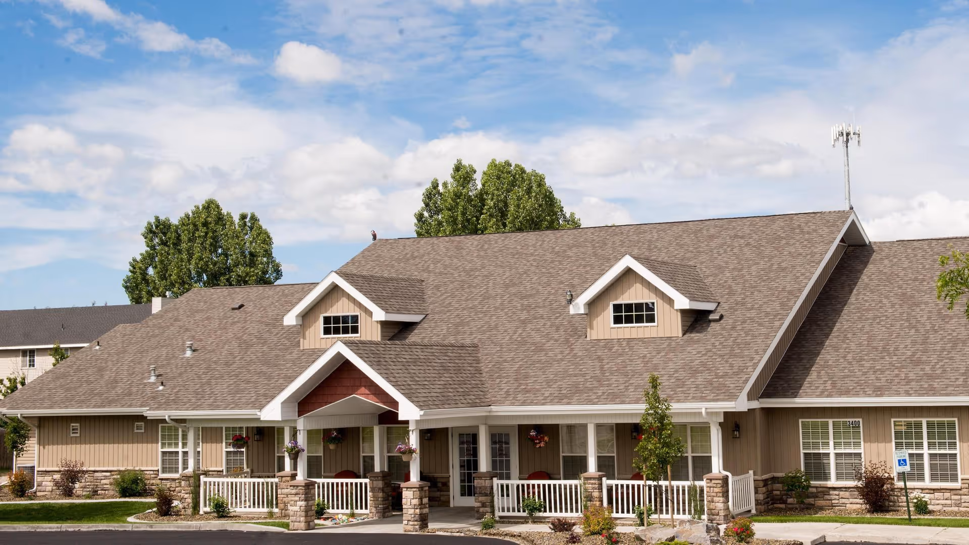 Exterior view of The Gables Assisted Living of Idaho Falls building with a large sloped roof, beige siding, stone accents, white trim, and a covered porch entrance. The building is surrounded by green grass, small trees, and shrubs under a partly cloudy blue sky.