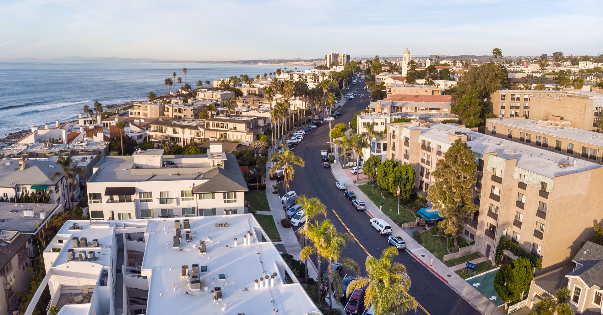 Aerial view of a coastal urban neighborhood with multiple residential buildings, palm trees lining the streets, and the ocean visible on the left side under a clear sky.