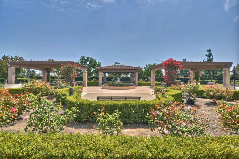 A well-maintained outdoor garden area with neatly trimmed hedges and blooming flowers. There are three wooden pergolas, one in the center with a fountain underneath and two on either side adorned with climbing plants. A bench is placed in front of the central pergola, and the sky is clear and blue.