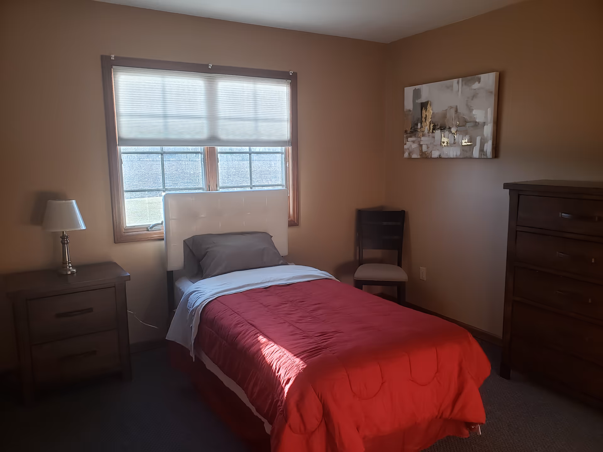 Single bed with red comforter, nightstands, dresser, and a window in a cozy bedroom.