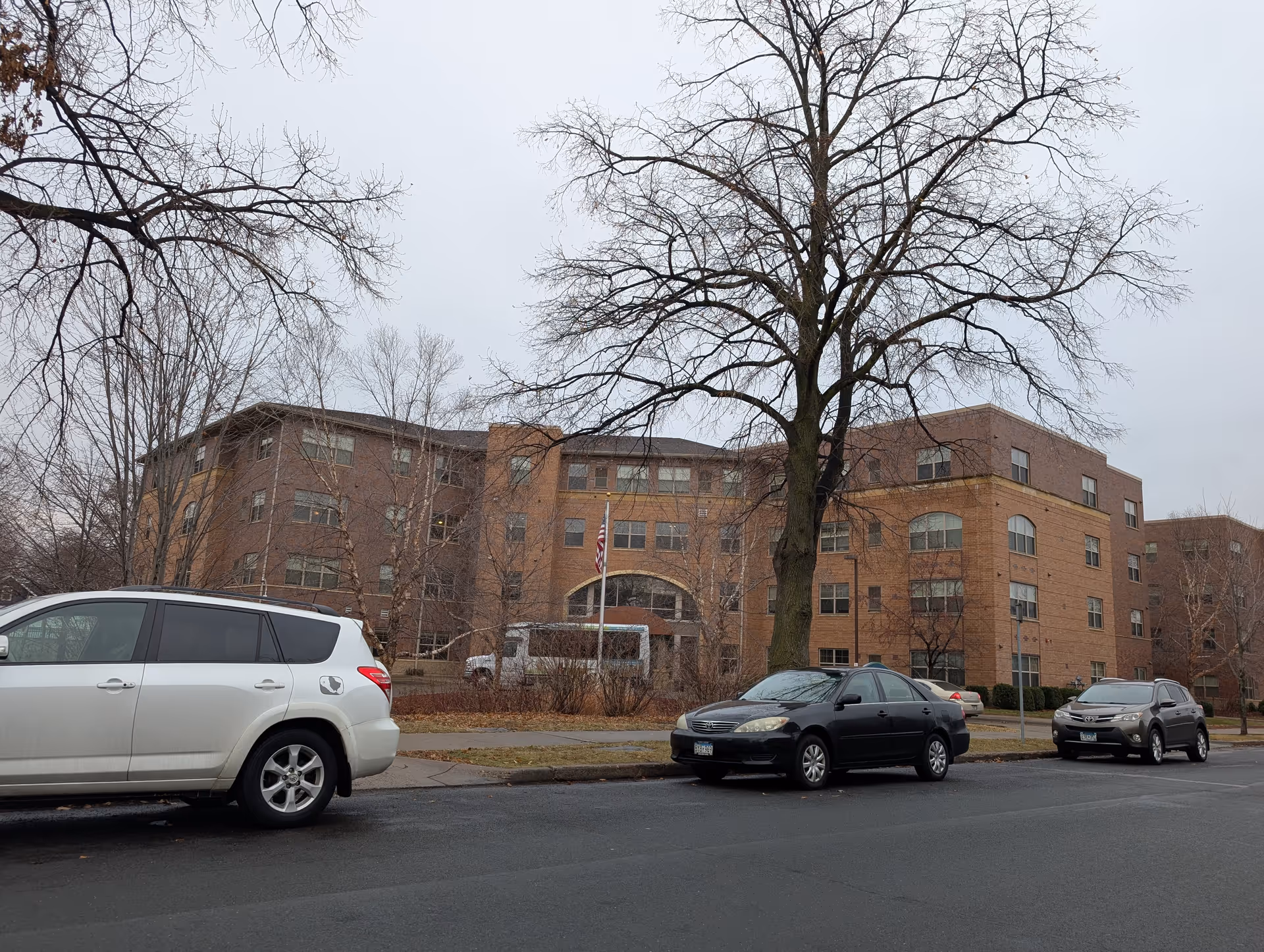 Exterior view of a multi-story brick building with several windows, an American flag in front, and a few parked cars along the street. Leafless trees are visible around the building under an overcast sky.