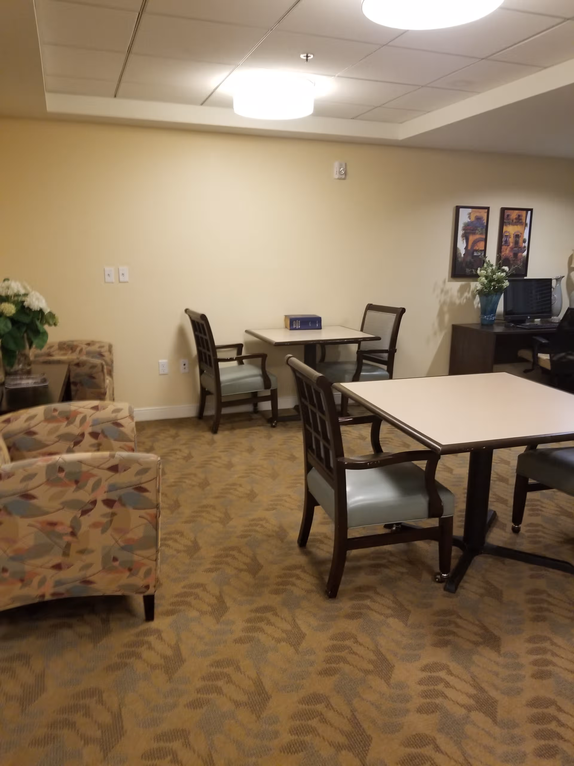 Cozy senior living common area with tables, chairs, sofa, and a desk in background.