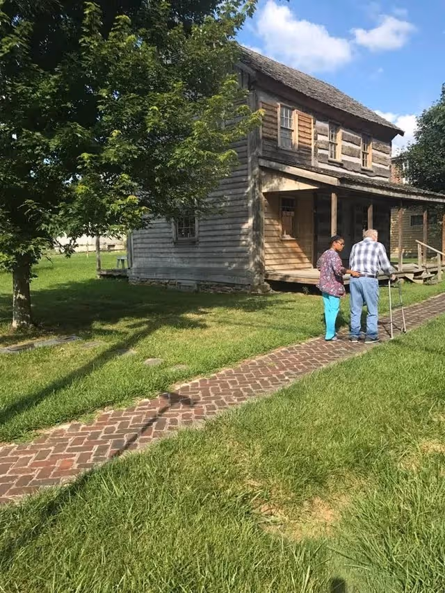 Two elderly residents stand with a walker near a weathered wooden building on a brick path.