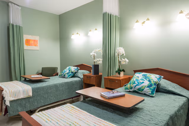 Twin-bed senior care room with green walls and bedding, a privacy curtain between beds, bedside tables with lamps and orchids, and over-bed tables.