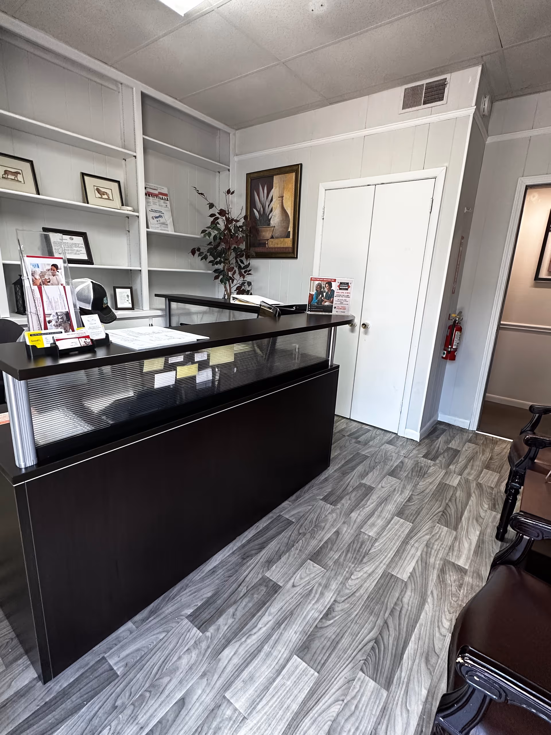 Reception area with a dark counter, shelves, plant, and framed art inside.