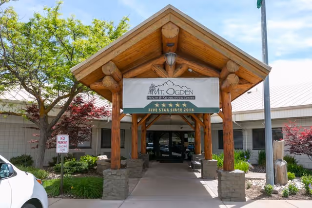 Covered log-beam entrance and driveway to Mt. Ogden Health and Rehabilitation Center with a sign and landscaped planting beds.