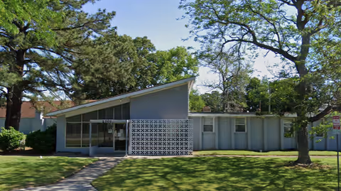 Single-story mid-century modern building with a slanted roof, decorative breeze-block wall and large front windows set behind a grassy lawn and trees.