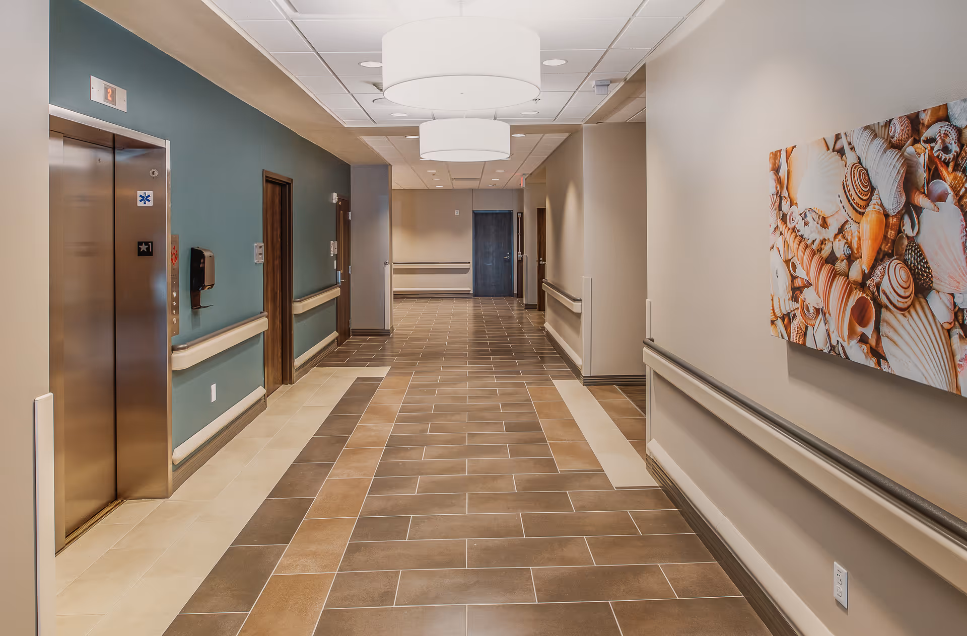 A clean, well-lit hospital hallway with tiled floors in shades of brown and beige. On the left side, there is a stainless steel elevator with a digital floor indicator showing the number 2. The walls are painted in a combination of teal and beige, with handrails along both sides of the corridor. Ceiling lights with large round fixtures illuminate the space. On the right wall, there is a colorful artwork featuring various seashells. Several closed doors line the hallway.