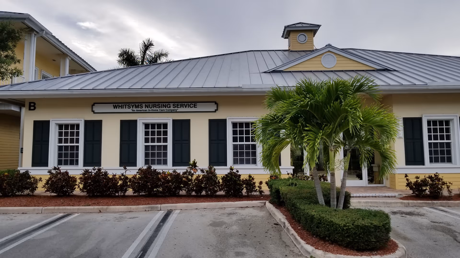 Front exterior of a beige building with green shutters and palm trees.
