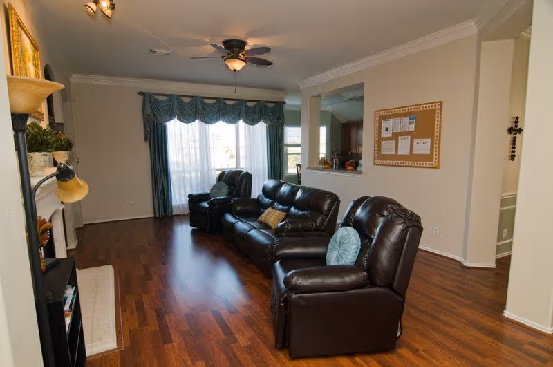 Living room with dark leather recliners and sofa on wood floors, a ceiling fan, large curtained window and a bulletin board on the wall.
